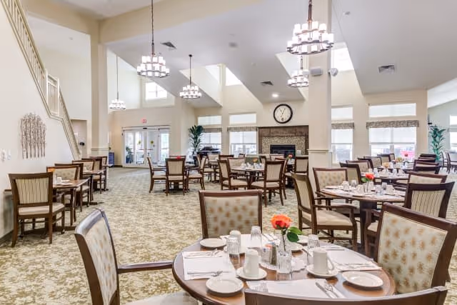 A spacious dining room with multiple round and square tables set with white plates, cups, glasses, and silverware. The room features high ceilings with hanging chandeliers, large windows allowing natural light, a fireplace with a clock above it, and carpeted floors with a floral pattern. The walls are painted light beige, and there are some plants and decorative elements around the room.