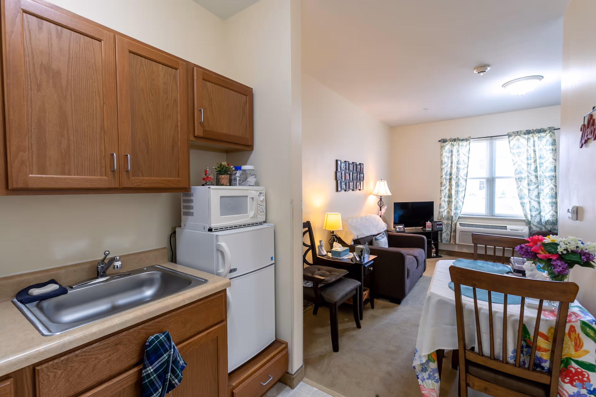 A small living space in a senior living facility featuring a kitchenette with wooden cabinets, a sink, a microwave, and a refrigerator. Adjacent to the kitchenette is a cozy living area with a brown armchair, a side table with a lamp, a TV on a stand, and a window with floral curtains. A dining table with a floral tablecloth and wooden chairs is also visible.