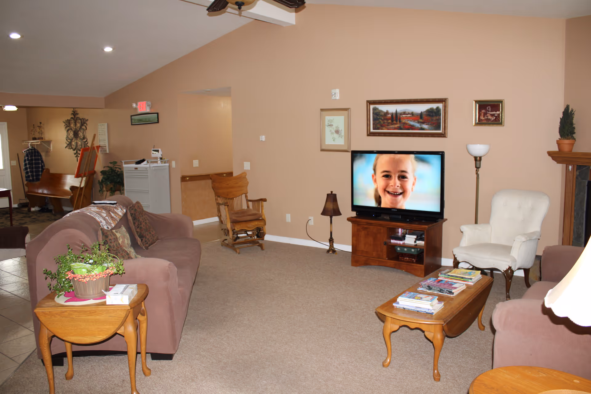 Comfortable communal living room with sofas, armchairs, coffee tables, a TV showing a child's face, and framed artwork on beige walls.