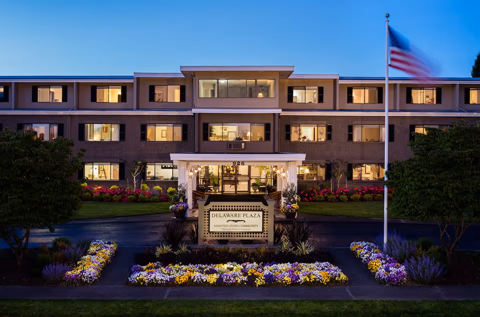 Exterior view of Delaware Plaza Assisted Living Community building at dusk with lights on inside. The three-story building has multiple windows, a central entrance with a covered porch, and a sign in front surrounded by colorful flower beds. An American flag on a flagpole is visible on the right side.