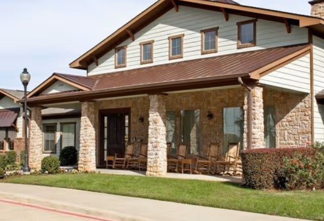 Exterior view of a single-story assisted living facility building with stone pillars, a brown metal roof, several windows, and a covered porch with rocking chairs. There is a well-maintained lawn and a streetlamp near the sidewalk.