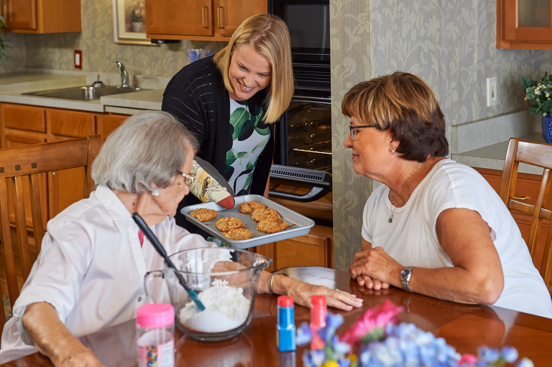 A woman wearing an oven mitt is taking a tray of freshly baked cookies out of the oven while two elderly women sit at a table watching and smiling. The table has a mixing bowl with flour and sugar, a spatula, and containers of sprinkles. The setting is a kitchen with wooden cabinets and a floral wallpaper.