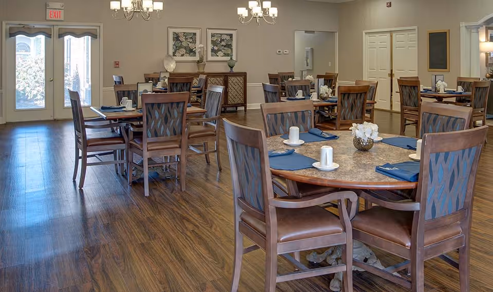 A senior living facility dining room with multiple round tables set with blue napkins, white cups, and small flower arrangements. The room has wooden floors, beige walls, and chandeliers hanging from the ceiling. There are double glass doors letting in natural light and framed floral artwork on the walls.