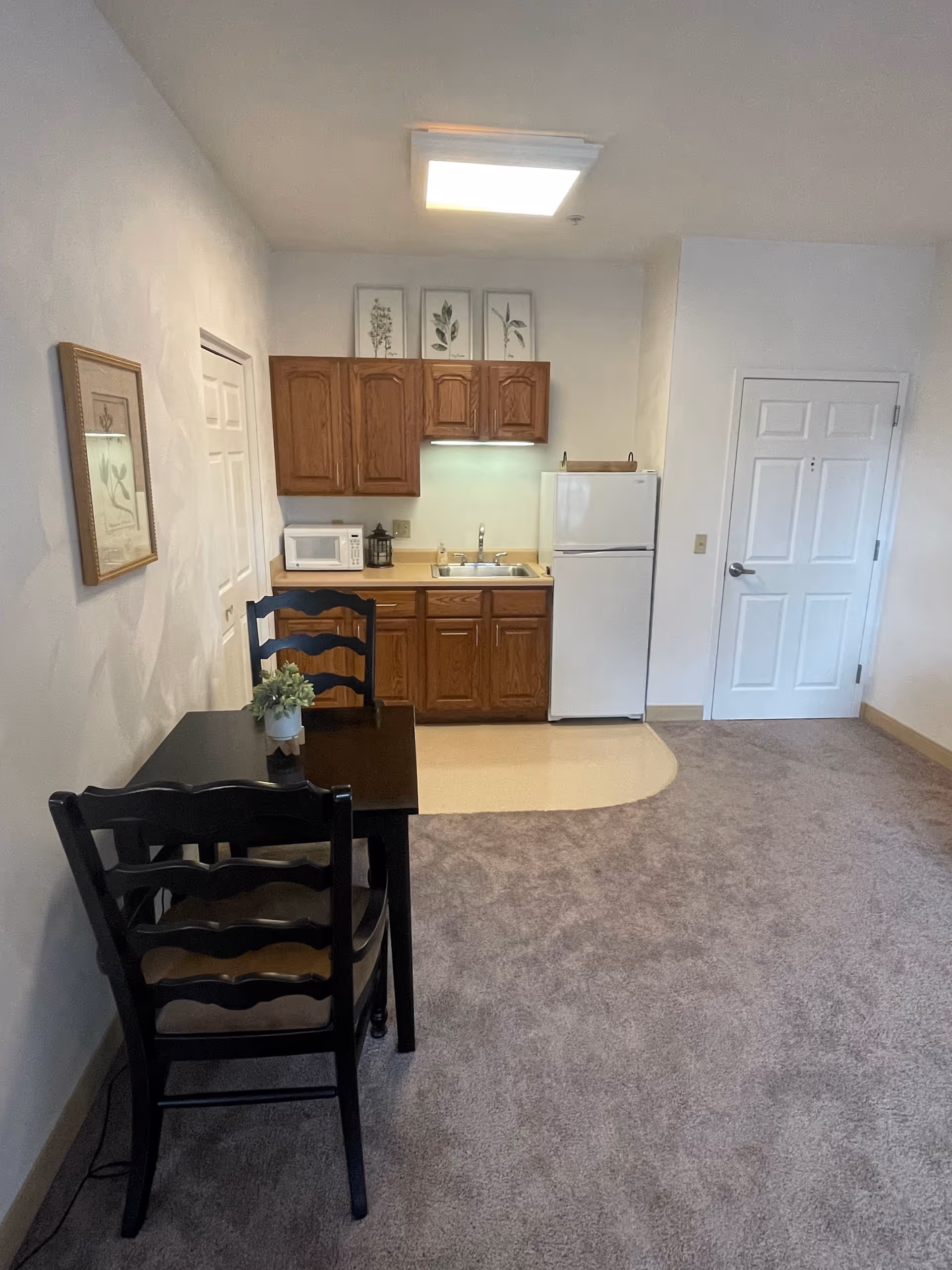 A small kitchenette area in a senior living facility with wooden cabinets, a white refrigerator, a microwave, and a sink. There is a small dining table with two black chairs and a small plant on the table. The floor is carpeted except for the kitchenette area which has a linoleum floor. The walls are white with framed botanical prints hanging above the cabinets and on the side wall. A white door is visible on the right side of the image.
