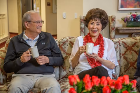 Two elderly people sitting on a floral patterned couch in a cozy living room, smiling and holding white mugs. There are red flowers in the foreground and framed artwork on the wall behind them.