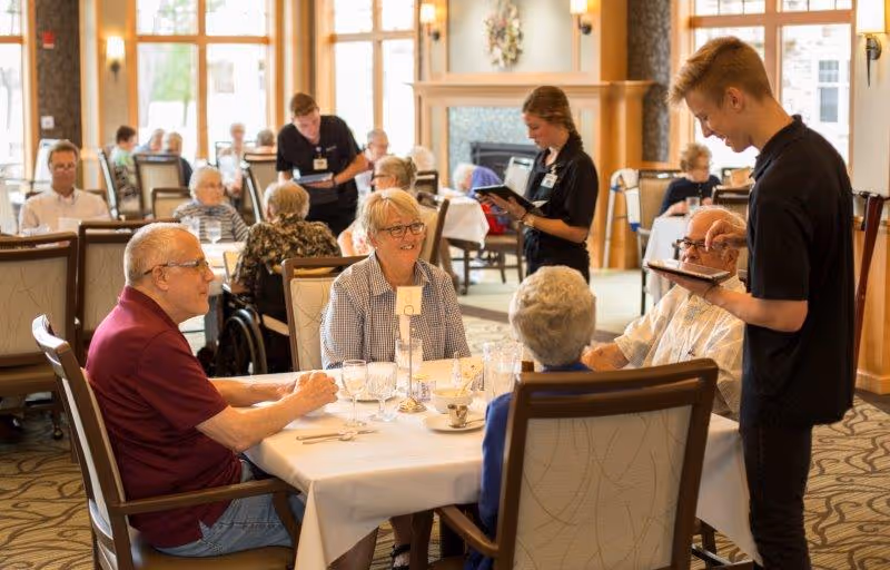 A group of elderly people seated around a dining table in a bright, spacious dining room with large windows. Two servers are attending to the guests, taking orders and serving food. The room has wooden accents and a cozy, welcoming atmosphere.