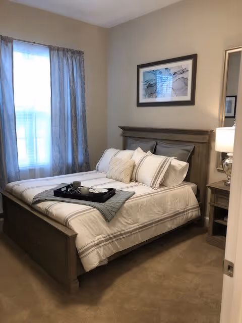A neatly made bed with white and beige bedding and multiple pillows in a bedroom. A black tray with a cup and plate is placed on the bed. There is a window with light blue curtains on the left wall, a framed abstract artwork above the bed, and a nightstand with a lamp and a mirror on the right side.