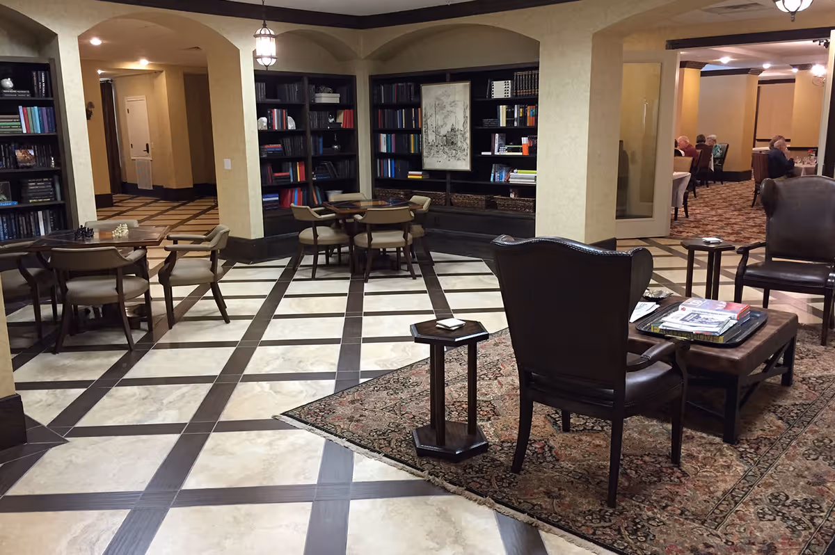 A cozy common area in a senior living facility featuring a tiled floor with dark wood accents, several wooden chairs and tables, a patterned rug, and built-in bookshelves filled with books. In the background, there is a dining area with elderly residents seated at tables. The walls are painted a warm beige color, and the space is softly lit with hanging lantern-style lights.