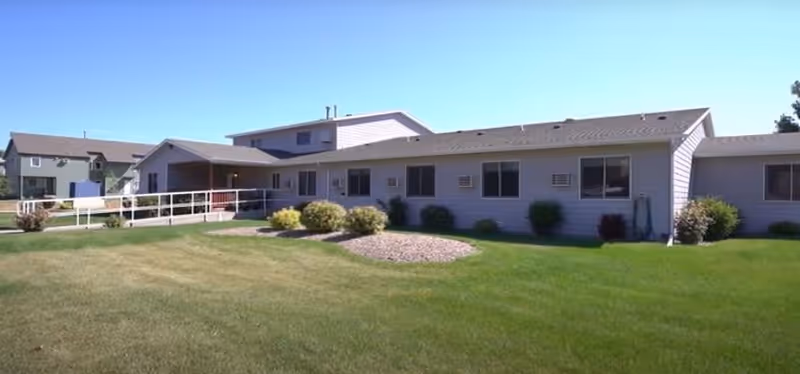 Exterior view of a single-story senior living facility building with a well-maintained green lawn and some shrubs under a clear blue sky.