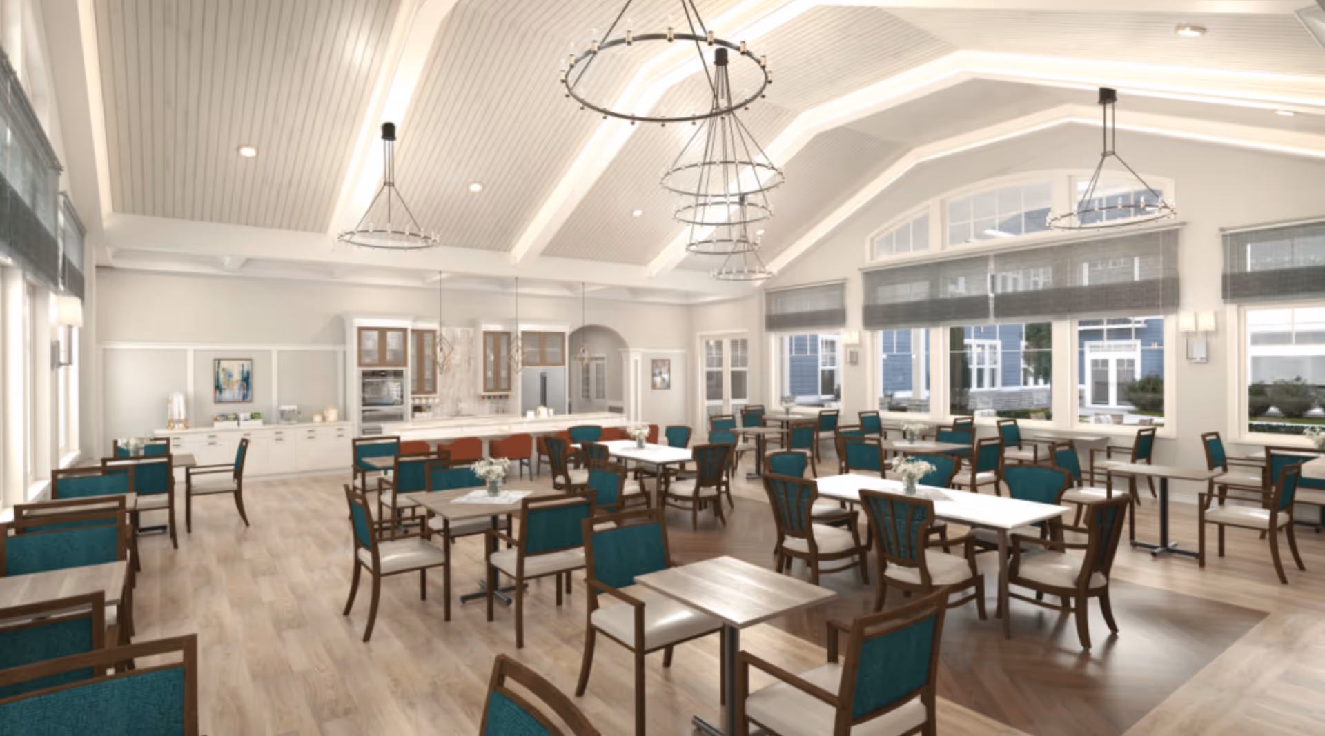 A spacious, well-lit dining room with multiple tables and chairs arranged neatly. The room features large windows with gray blinds, a high vaulted ceiling with modern circular chandeliers, and a kitchen area with white cabinetry and a marble backsplash in the background.