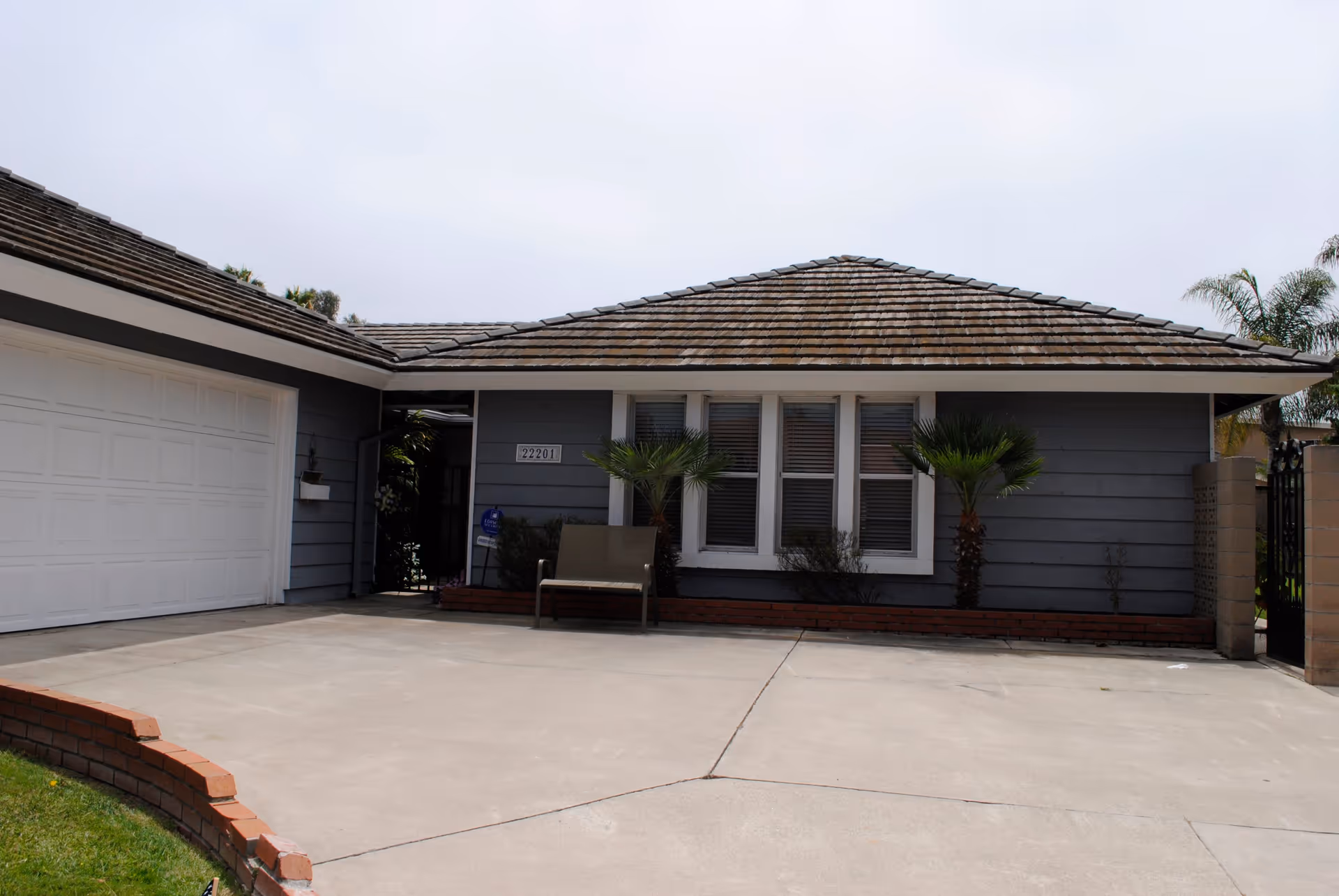Front exterior view of a single-story house with a gray exterior, a tiled roof, a white garage door on the left, three windows with white frames in the center, two small palm trees and some shrubs in front of the windows, a bench near the entrance, and a concrete driveway.