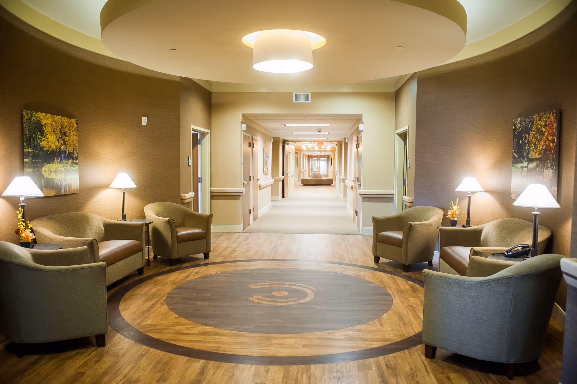 A well-lit seating area in a hallway of a senior living facility with six armchairs arranged around small tables with lamps and floral decorations. The walls are painted in warm tones and decorated with framed nature artwork. The hallway extends into the distance with multiple doors on either side.