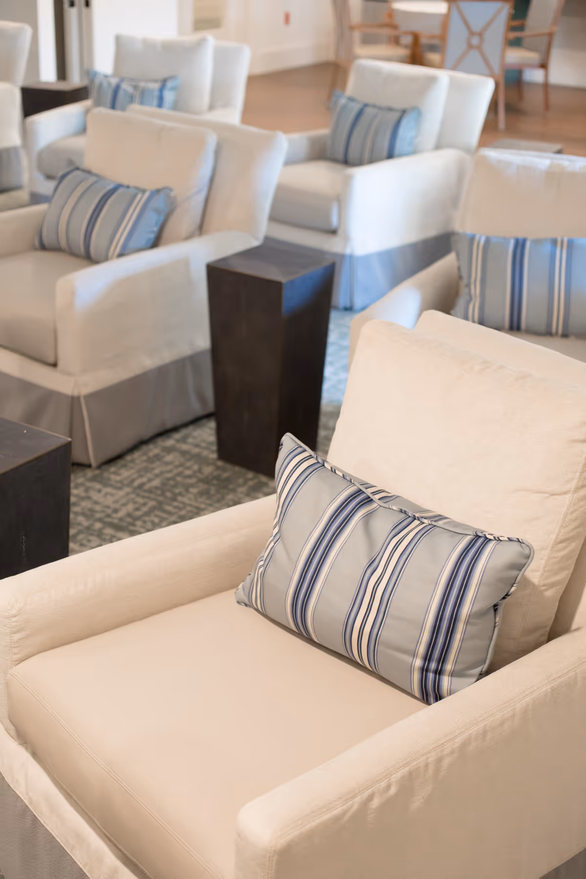A cozy seating area with several beige armchairs, each adorned with a blue and white striped pillow. The chairs are arranged on a patterned carpet with small dark wooden side tables between them. The background shows more seating and part of a wooden table with chairs.