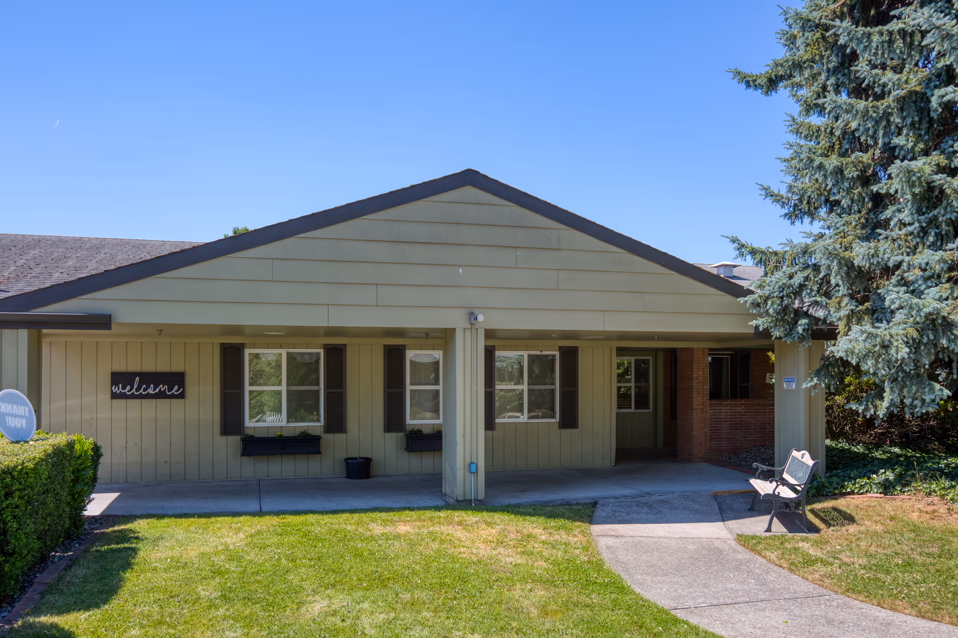 Exterior view of a single-story building with beige siding and a dark roof. The building has two windows with black shutters and flower boxes, a covered entrance area with a bench on the right side, and a 'welcome' sign on the left wall. There is a well-maintained lawn and a concrete walkway leading to the entrance. A large evergreen tree is visible on the right side under a clear blue sky.