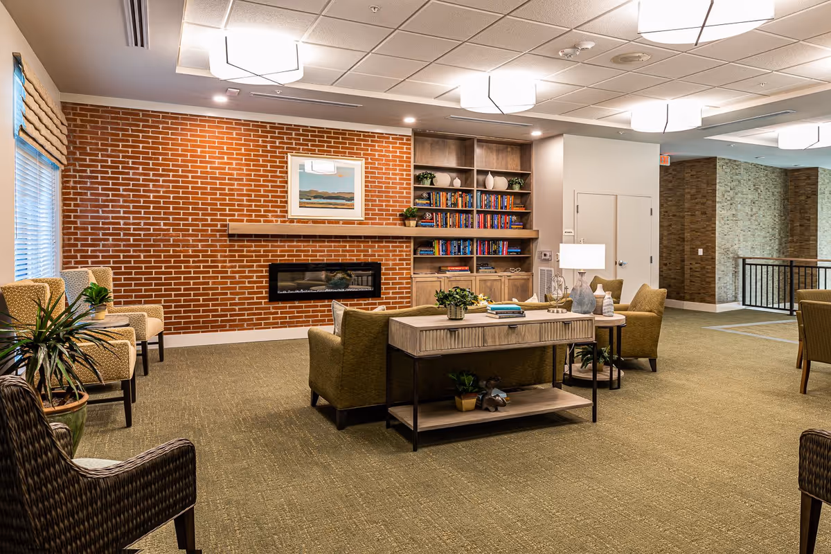 A cozy living room area with a brick accent wall featuring a modern electric fireplace and a framed landscape painting above it. There is a built-in wooden bookshelf filled with books and decorative items. The room has several upholstered chairs and a sofa arranged around a wooden console table with plants and books. The space is well-lit with ceiling lights and has carpeted flooring.