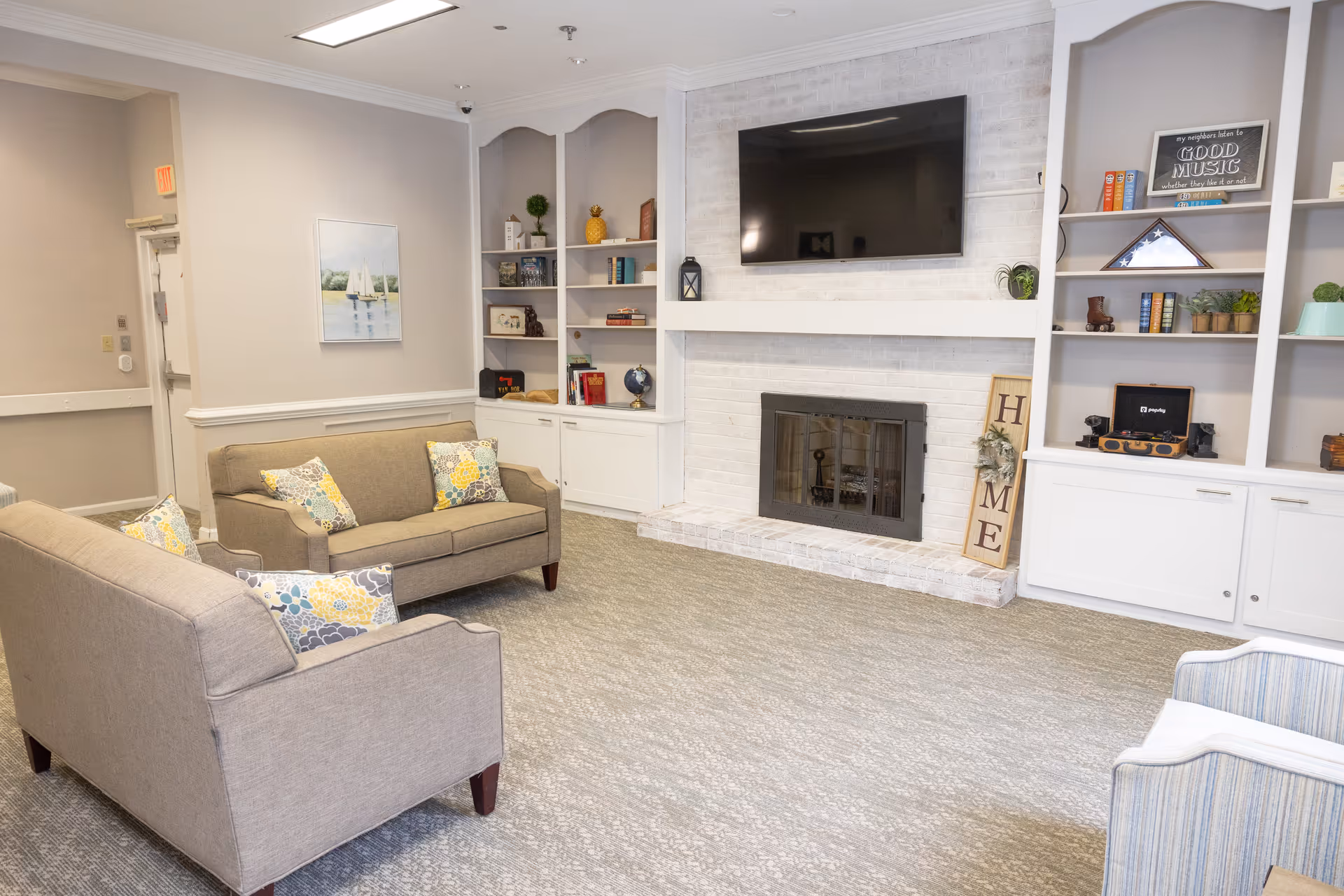 A cozy living room area in a senior living facility featuring two beige sofas with patterned cushions, a white brick fireplace with a mounted flat-screen TV above it, built-in white shelves with decorative items and books, and a light gray carpeted floor.