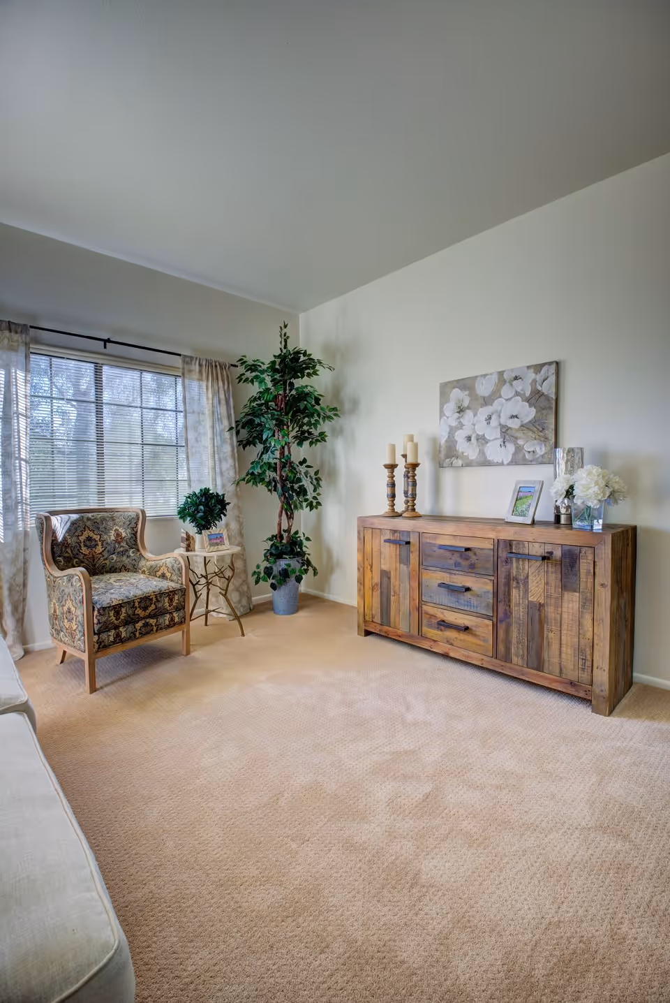A cozy living room area with beige carpet, a wooden sideboard decorated with candles, a framed picture, and a vase of white flowers. There is a patterned armchair next to a small round table with a plant on it, and a tall potted plant near a window with sheer curtains allowing natural light to enter.