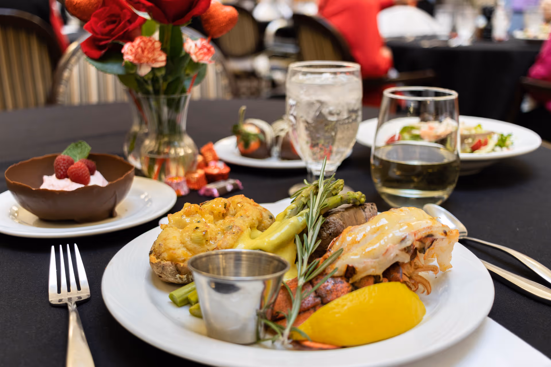 Close-up of a plated upscale meal with steak, asparagus, a stuffed potato, a lemon wedge and dessert on a black tablecloth in a dining setting.
