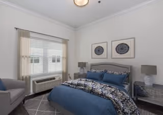 Well-lit bedroom with a blue bed, nightstands and lamps, an armchair, and a curtained window with an AC unit.