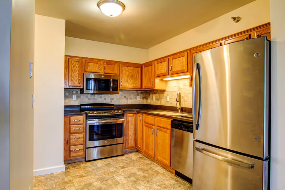 A modern kitchen with wooden cabinets, stainless steel appliances including a refrigerator, oven, microwave, and dishwasher. The kitchen has a tiled backsplash and a single overhead light fixture.