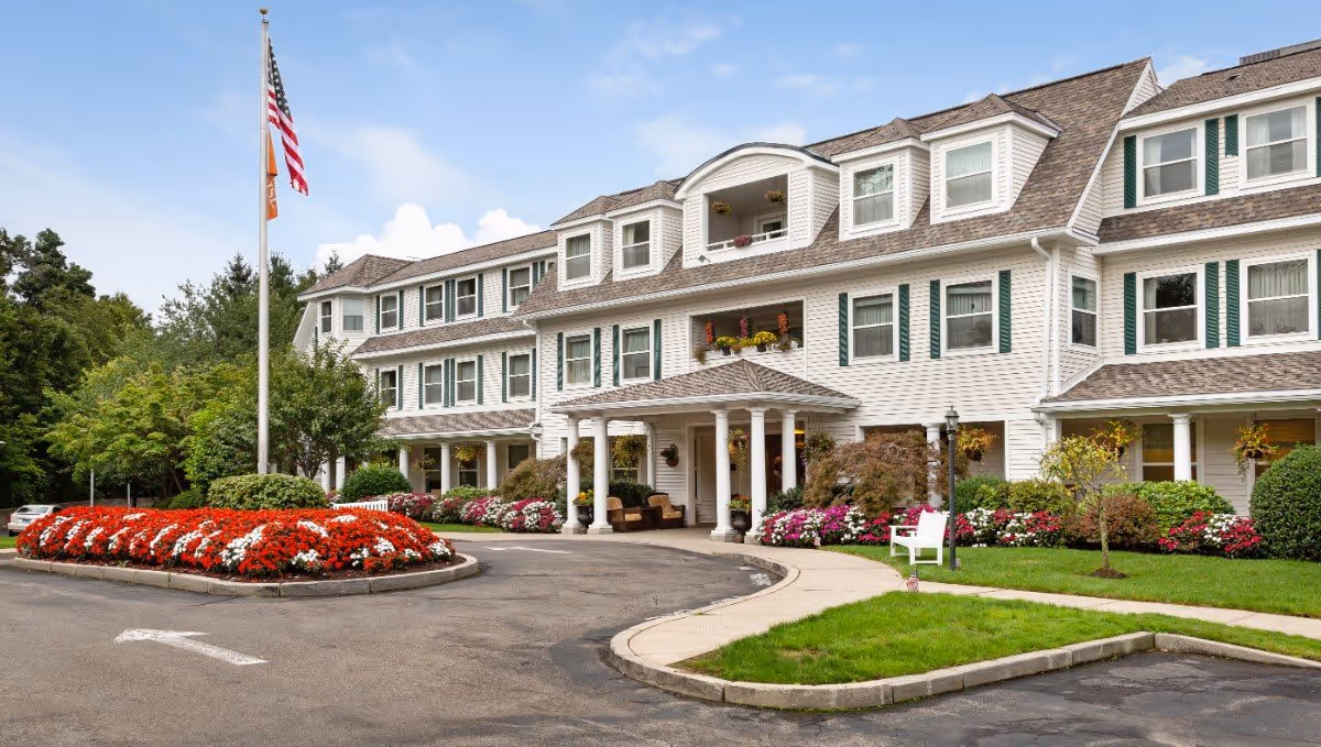 Exterior front view of a senior living facility with white siding and green shutters. The building has multiple windows and a covered entrance with columns. There is a circular driveway with a landscaped island featuring red and white flowers, an American flag on a flagpole, and well-maintained greenery and bushes around the building.
