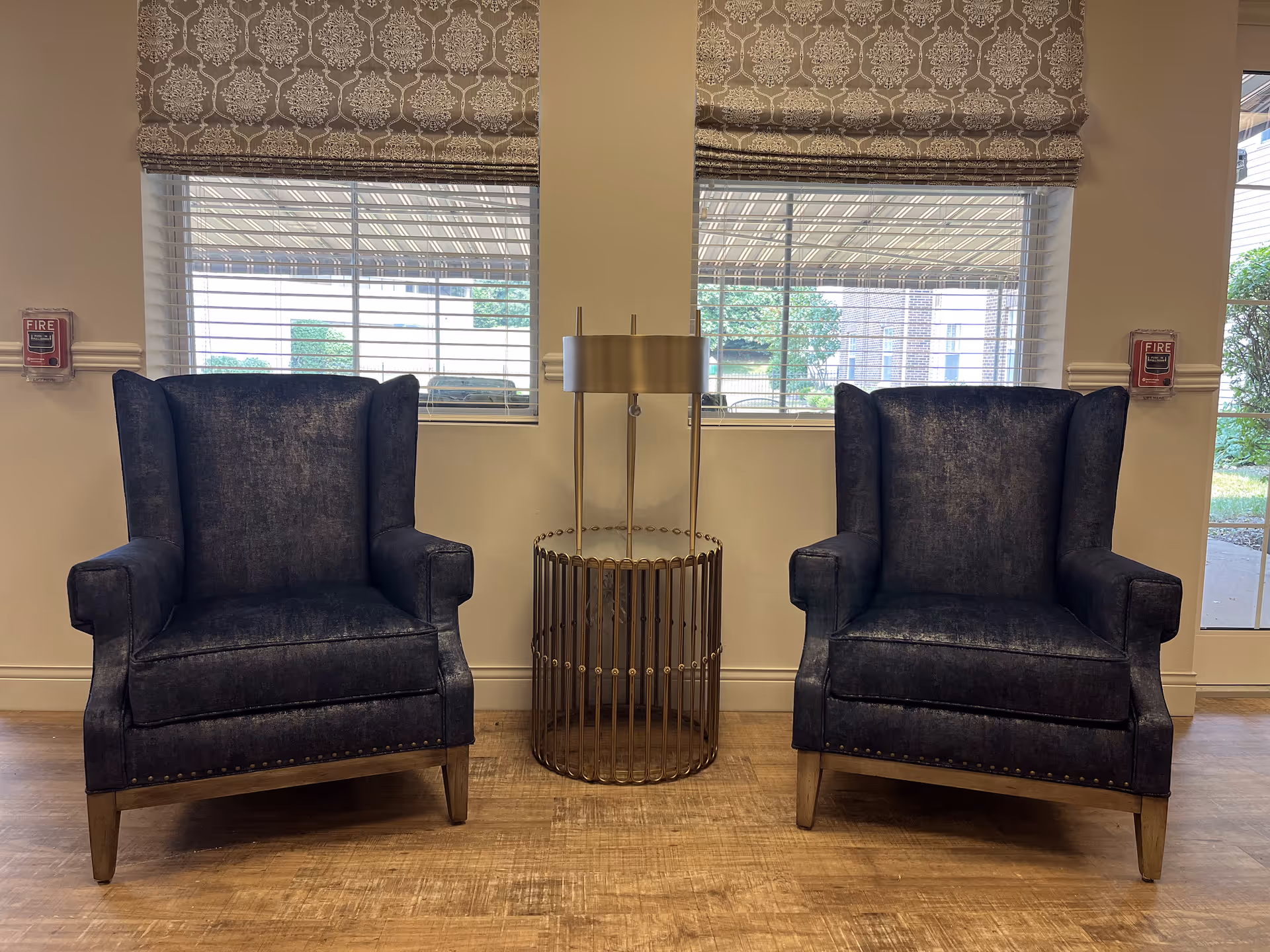 Two dark blue upholstered armchairs with wooden legs are positioned on either side of a small round gold metal side table with a modern gold lamp on top. Behind the chairs are two windows with white blinds and patterned Roman shades. The room has light-colored walls and wood flooring.