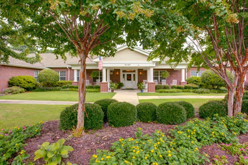 Front exterior view of a single-story assisted living facility with a brick facade, white columns, and a covered entrance. The building is surrounded by well-maintained landscaping including trimmed bushes, trees, and flowering plants. A concrete walkway leads to the entrance.