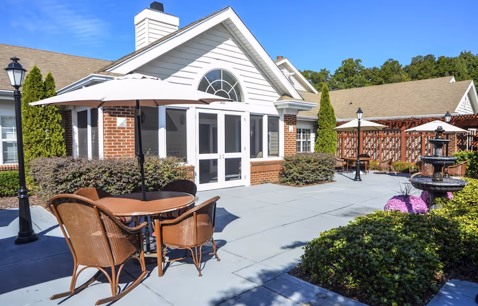 Outdoor patio area at Legacy Heights Senior Living Community with round tables and chairs under large umbrellas, surrounded by greenery, a water fountain, and a brick and white building under a clear blue sky.