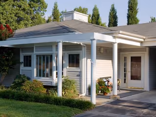 Entrance facade of a single-story senior living building with a covered porch supported by white columns, windows with blinds, and landscaped greenery.