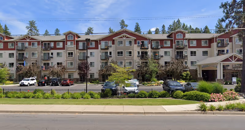 Exterior view of a multi-story senior living facility named Affinity at Mill Road with balconies, parked cars in front, landscaped greenery, and a covered entrance on the right side.