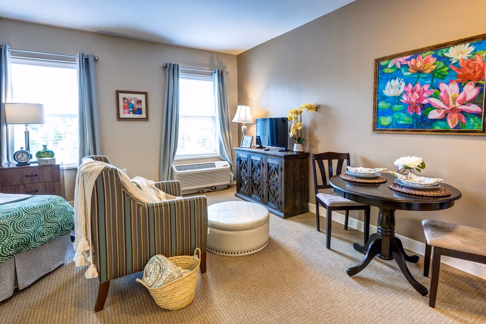 A cozy living area in a senior living facility with a striped armchair, a round ottoman, a small round dining table set for two, a wooden cabinet with a TV on top, two windows with light blue curtains, a colorful floral painting on the wall, and a bed partially visible with a green patterned blanket.
