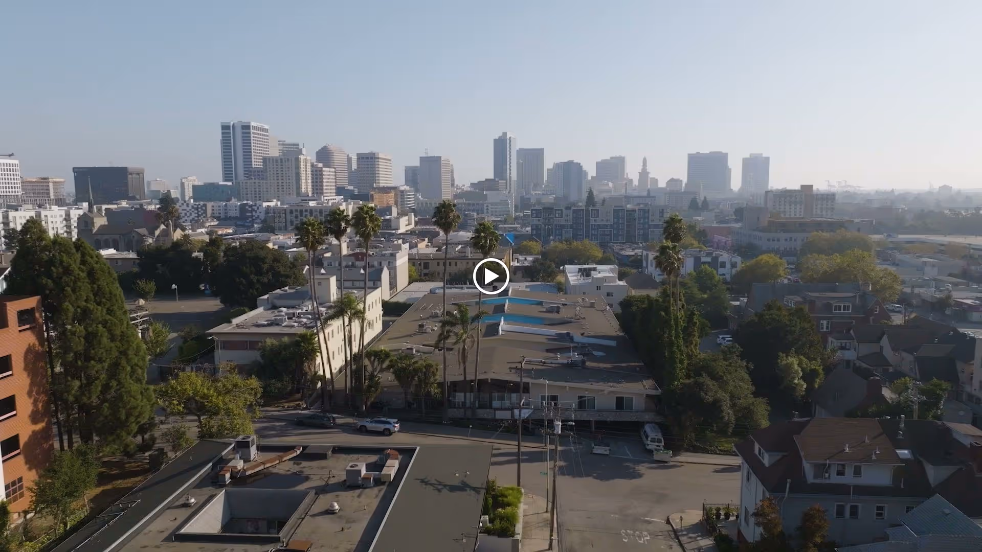 Aerial view of a city neighborhood with low-rise buildings, palm trees, and a distant downtown skyline.