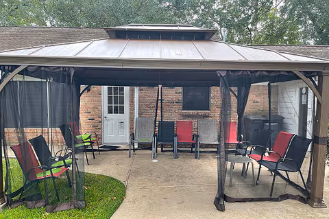 Outdoor seating area under a gazebo with mesh curtains, featuring several chairs with red and black cushions arranged on a concrete patio in front of a brick building with a white door and window.
