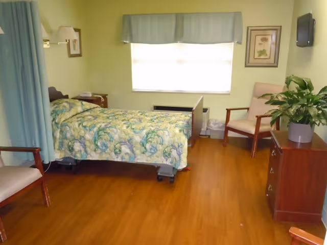 A simple bedroom in a care center with a single bed covered in a floral patterned quilt, a wooden nightstand with a lamp and telephone, two wooden chairs with cushions, a wooden dresser with a potted plant, a wall-mounted TV, a window with a valance, and light green walls.