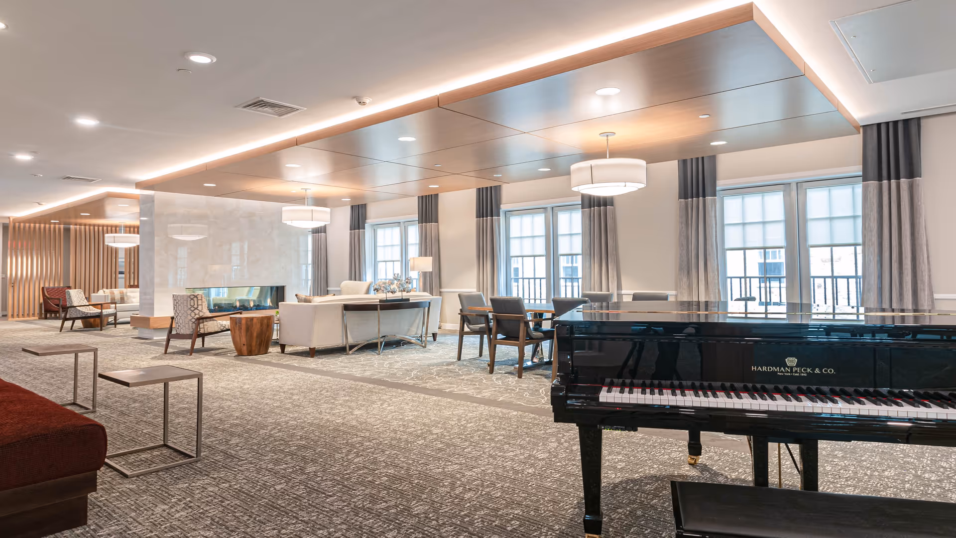 A spacious and well-lit common area in a senior living facility featuring a black grand piano in the foreground, several seating arrangements with armchairs and sofas around a modern fireplace, large windows with curtains allowing natural light, and contemporary ceiling lights.