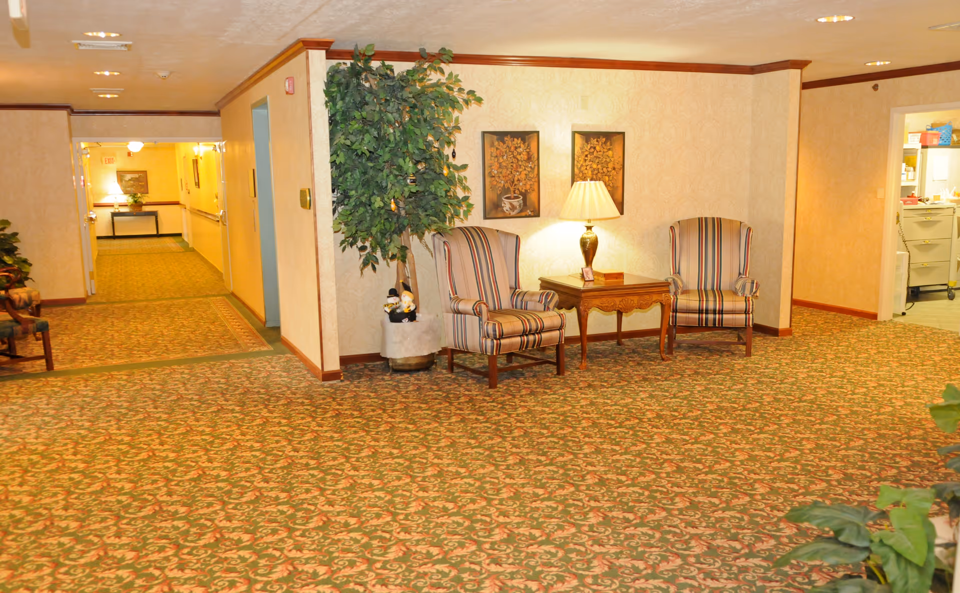 A cozy interior hallway area with patterned carpet and wallpaper. Two striped armchairs flank a wooden side table with a lamp and decorative items. A large potted plant is beside the chairs. The hallway extends to the left with more seating and artwork visible. A doorway on the right leads to a room with filing cabinets and office supplies.