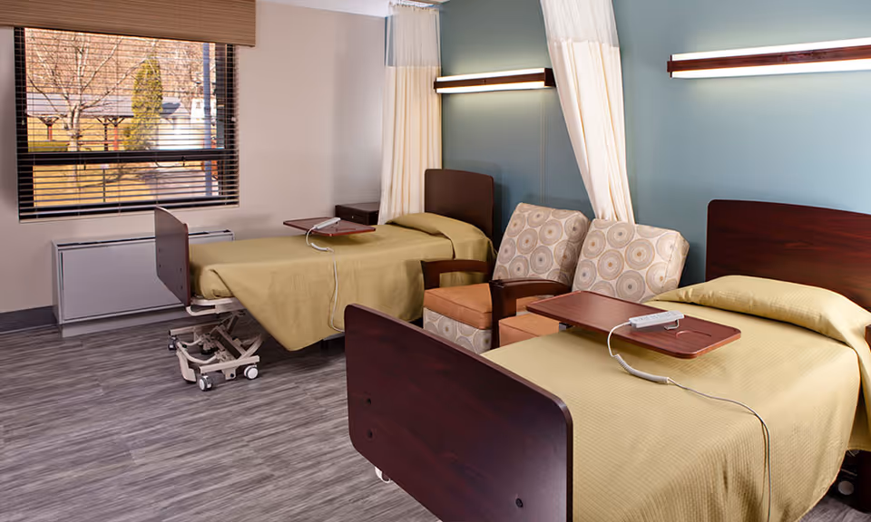 A rehabilitation room with two hospital beds covered in beige blankets, each equipped with an overbed table and a remote control. Between the beds are two patterned armchairs and a small side table. The room has a large window with blinds, light blue walls, and modern lighting fixtures above each bed.
