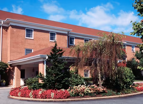 Exterior view of a three-story brick building with a covered entrance, surrounded by landscaped greenery including bushes, flowering plants, and trees under a blue sky with some clouds.