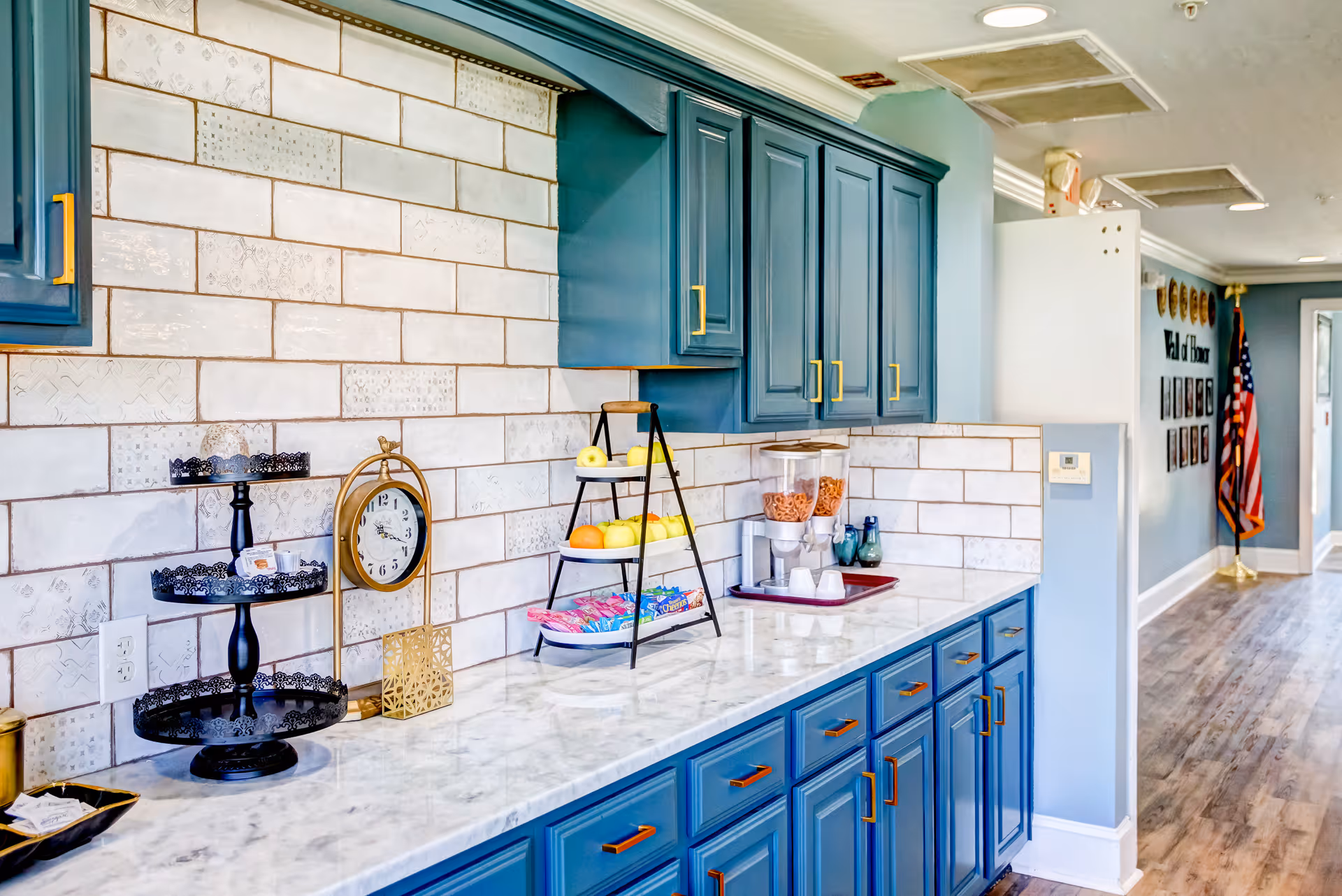 A bright kitchen area with blue cabinets and drawers featuring gold handles. The countertop is white marble with a three-tier black metal stand, a round clock, a tiered fruit stand with apples and oranges, and a cereal dispenser with cups on a tray. The backsplash consists of white tiles with subtle decorative patterns. In the background, a hallway with wooden flooring leads to a wall with framed photos and an American flag.