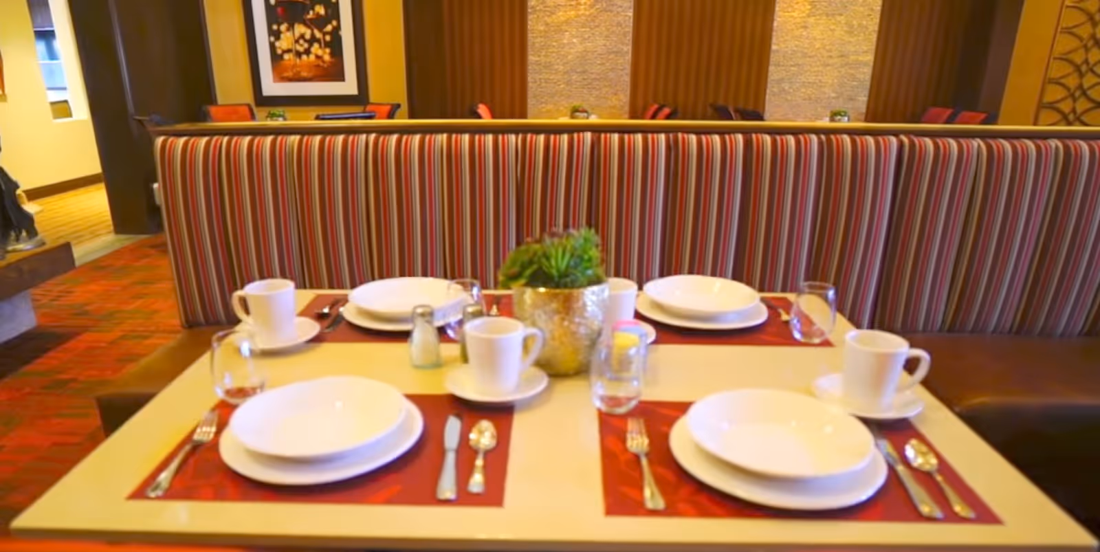 Table set for four with white dishes, cups, and cutlery on placemats in front of a striped booth in a dining area.