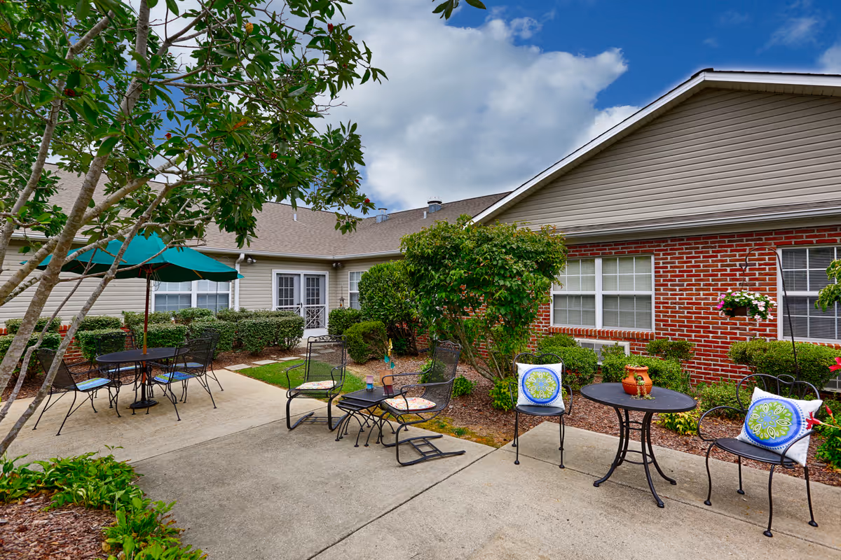 Outdoor patio area at Columbia Cottage Florence featuring metal chairs with colorful cushions, round tables, a green umbrella, and surrounding greenery including bushes and trees, with a building in the background under a partly cloudy sky.