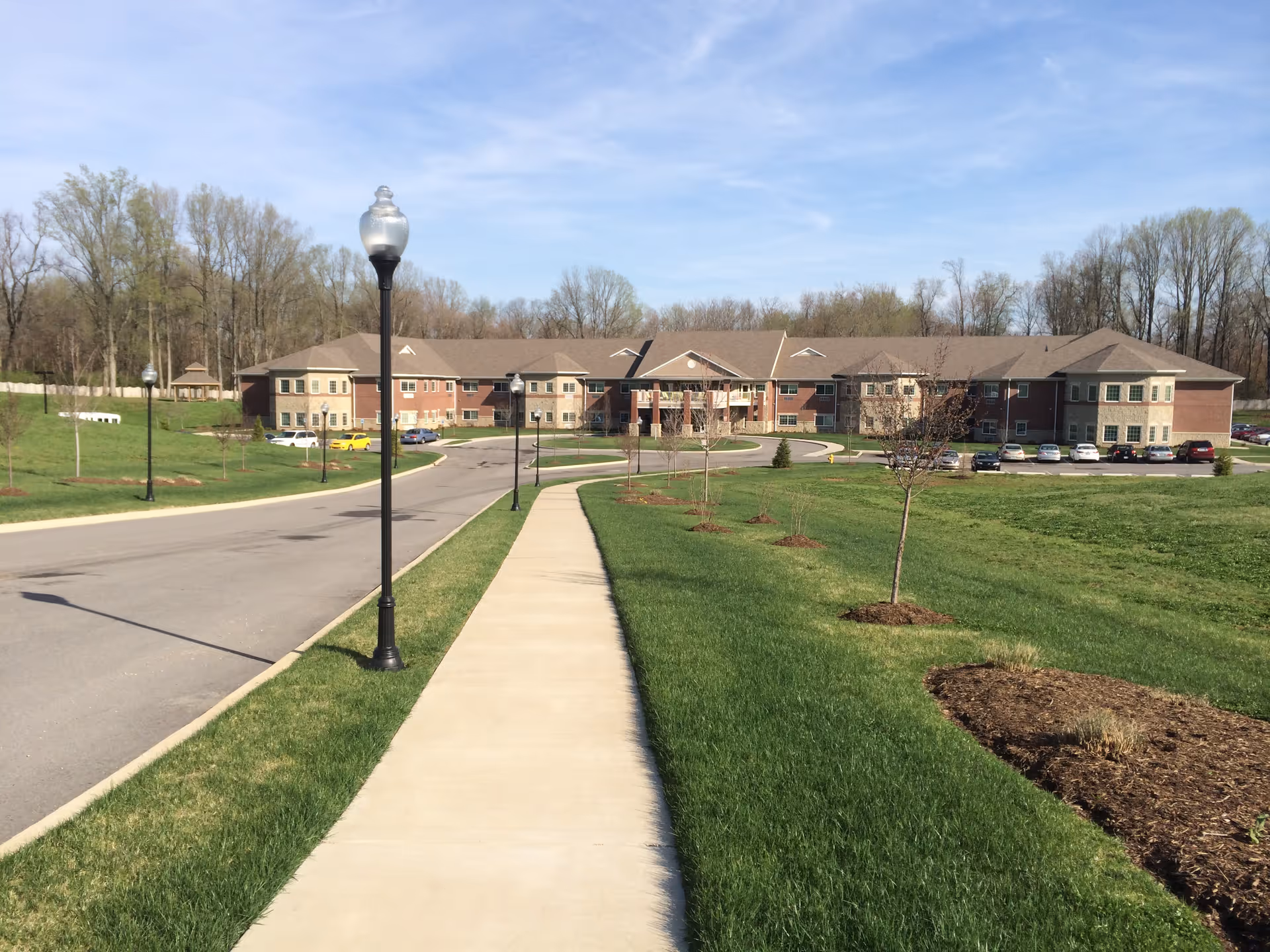 Concrete sidewalk flanked by lamp posts and green lawns leading to the front of a large two-story senior living building.