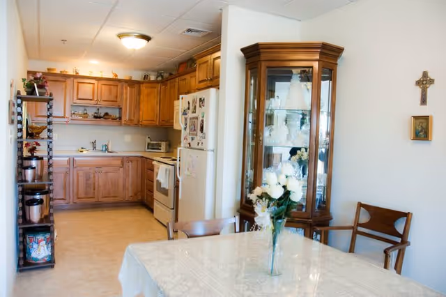 A cozy kitchen and dining area with wooden cabinets, a white refrigerator covered with magnets and photos, a stove, and a microwave. A dining table with a white tablecloth and a vase of white flowers is in the foreground. There is a wooden china cabinet with glass doors displaying decorative items, and a wooden chair against the wall with a cross and a small framed picture hanging above it.