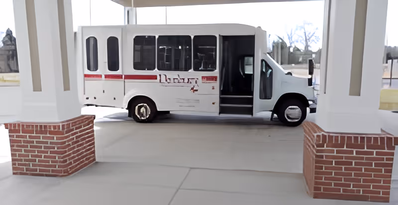A white shuttle bus parked under a covered drop-off area with brick pillars at a senior living facility.
