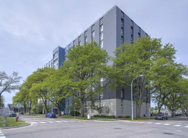 Exterior view of a multi-story residential building with gray and blue panels, surrounded by green trees along the street on a cloudy day.