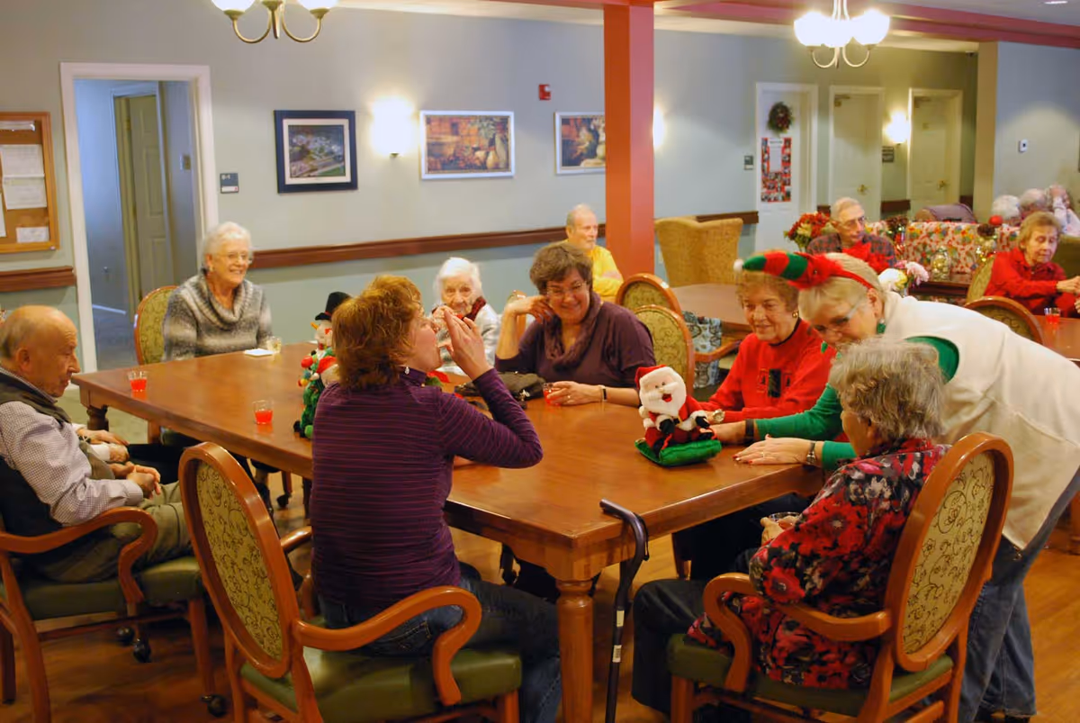 A group of elderly people sitting around a large wooden table in a common room, engaging in conversation and activities. One person is wearing festive holiday attire with a red and green elf hat, and there are holiday decorations on the table and walls. The room has warm lighting and framed pictures on the walls.
