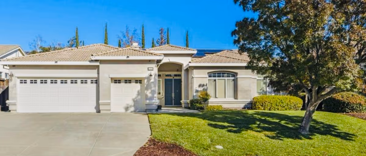 Front exterior view of a single-story house with a three-car garage, a tiled roof, a double front door, and a well-maintained lawn with a tree and shrubs under a clear blue sky.