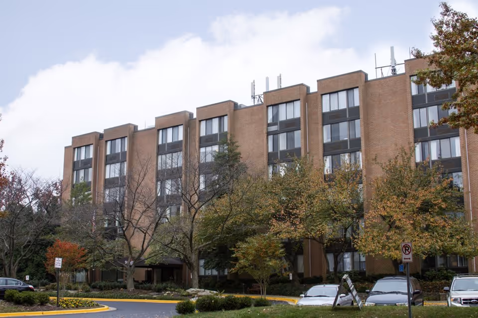 Exterior view of a multi-story brick building with large windows, surrounded by trees with autumn foliage and a parking area with several cars in front.