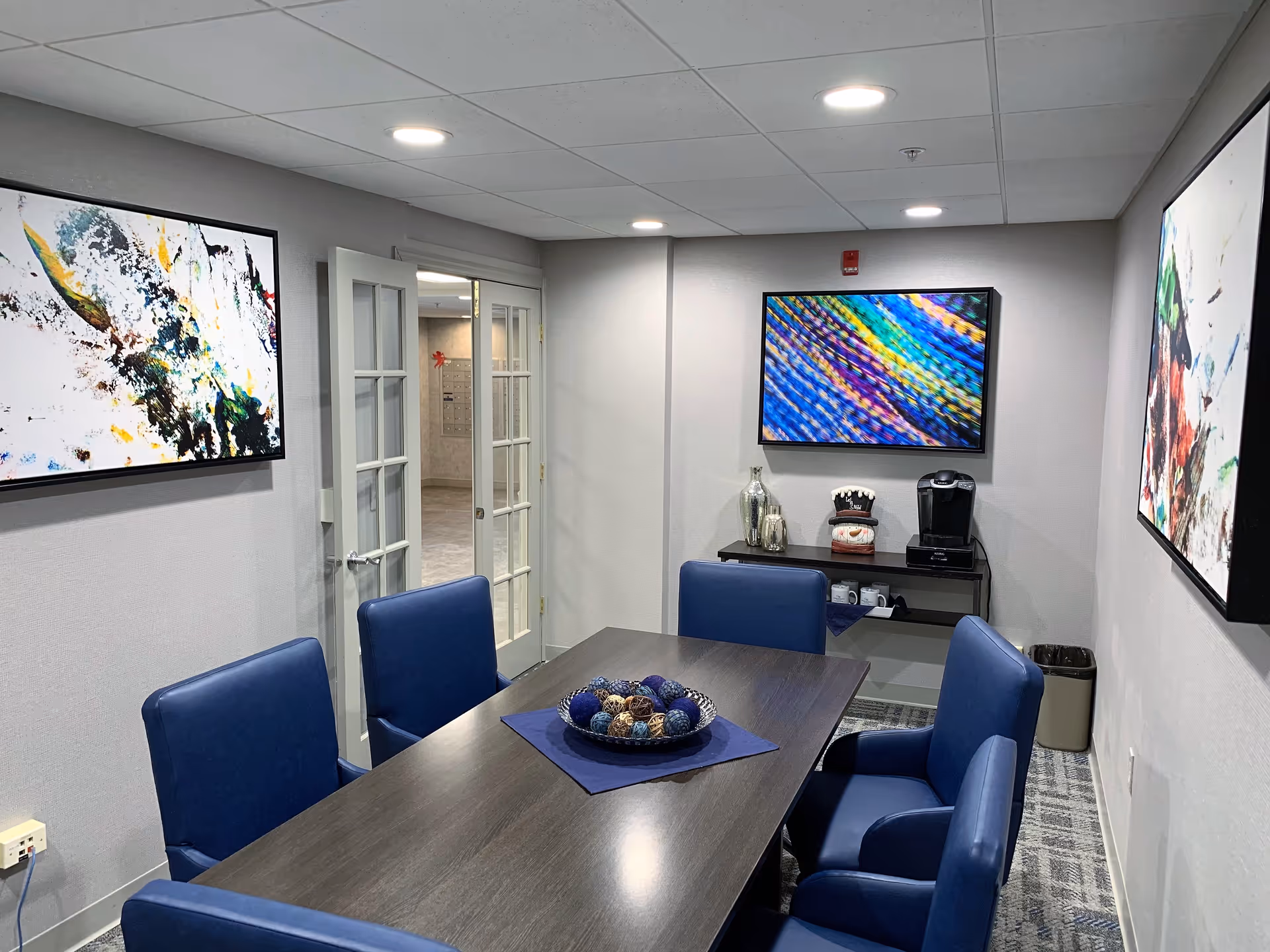Small interior meeting/dining room with a dark table, blue chairs, colorful wall art, and a coffee station.
