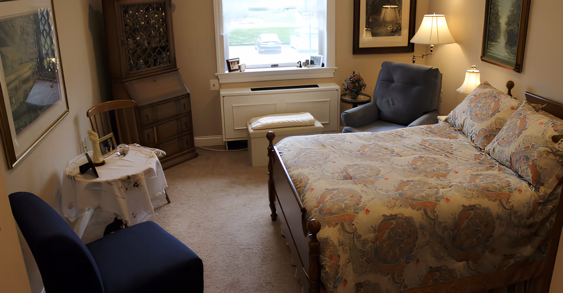 A cozy furnished bedroom with a patterned double bed, upholstered chair, small table, dresser and a window letting in daylight.